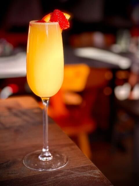 A tall champagne flute filled with a yellow-orange Apple Cider Mimosas cocktail, garnished with a slice of orange and a strawberry, sits on a wooden table with a blurred background.