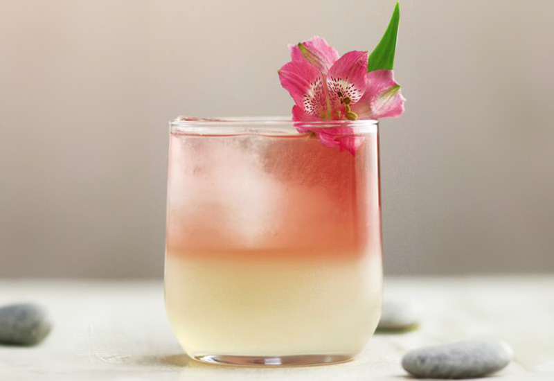 A two-toned cocktail in a clear glass, with a pale yellow layer on the bottom and a pink layer on top, garnished with a pink flower and green leaf. Smooth stones are blurred in the background.
