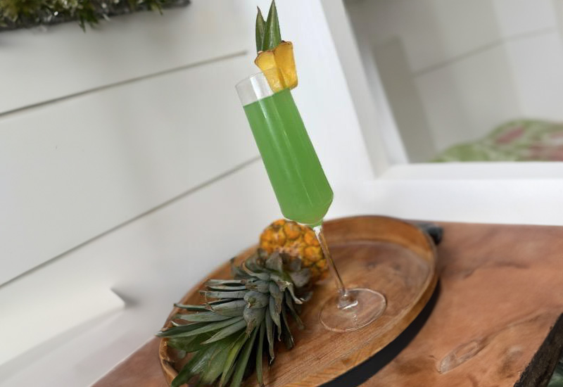 A tall glass of green cocktail garnished with pineapple wedges sits on a wooden tray, next to a whole pineapple, against a white background.