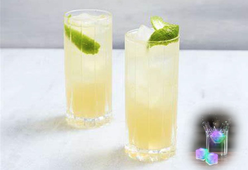 Two tall glasses filled with a light yellow drink and ice, each garnished with a lime wedge, sit on a white surface. In the lower right corner, a small, glowing cocktail with colorful ice cubes is shown.