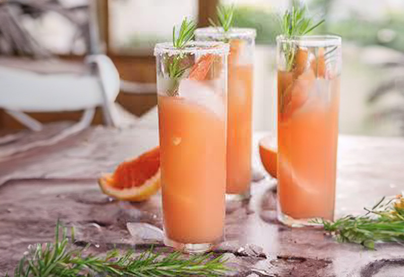 Three tall glasses of pink grapefruit cocktail with ice, garnished with rosemary sprigs and a salt rim. Slices of grapefruit and rosemary are scattered on the table in a bright, casual setting.