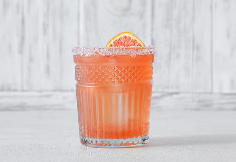A pink cocktail in a textured glass, garnished with a grapefruit slice and a sugared rim, sits on a light surface with a white wooden background.