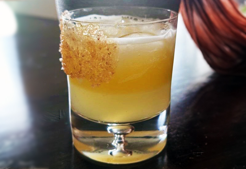 A yellow cocktail with a frothy top is served in a short glass, garnished with a crisp, golden sugar shard on the rim. The drink sits on a dark surface with a blurred background.