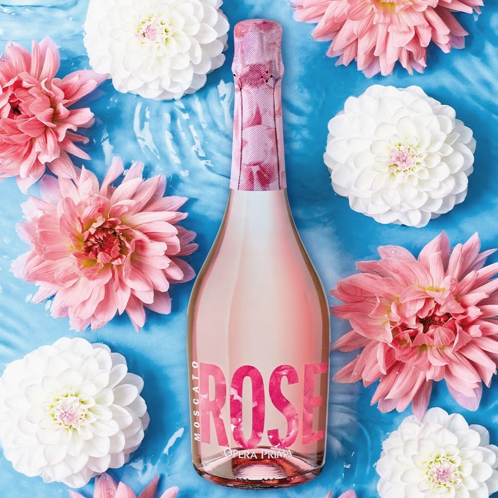 A bottle of rosé Moscato wine with a pink label is surrounded by pink and white flowers on a light blue watery surface.