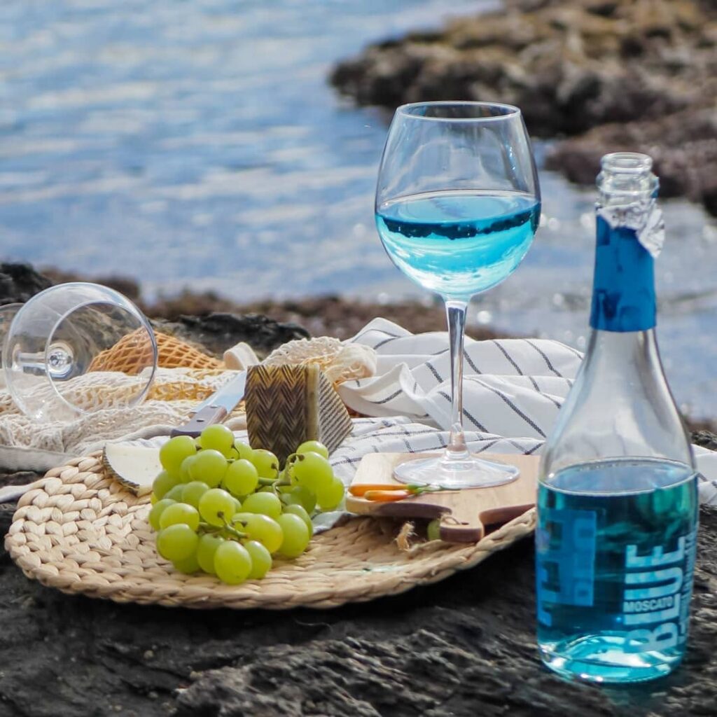 A picnic by the water with a glass and bottle of blue Moscato, green grapes, cheese, and bread on a woven mat, with a glass and sea in the background.
